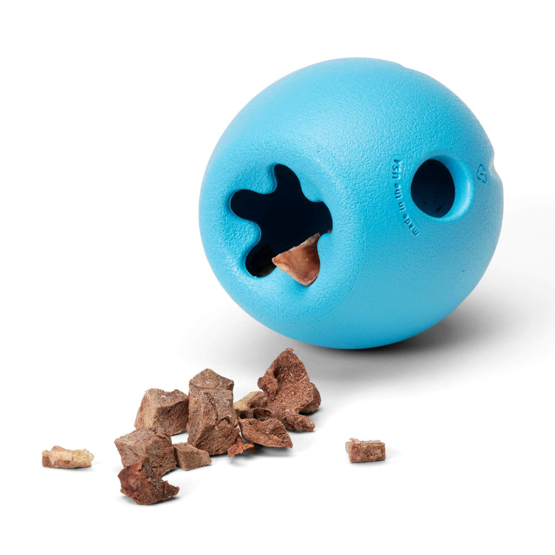 A bright blue textured rubber dog toy ball with a star-shaped hole and a circular hole, shown on a white background with brown kibble pieces spilled in front.