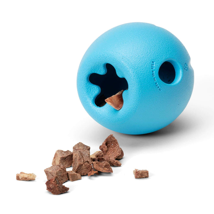 A bright blue textured rubber dog toy ball with a star-shaped hole and a circular hole, shown on a white background with brown kibble pieces spilled in front.