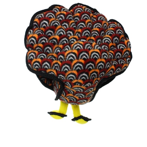 Rear view of a plush turkey-shaped dog toy, showing colorful feather patterns in black, orange, and white, with yellow feet visible at the bottom.